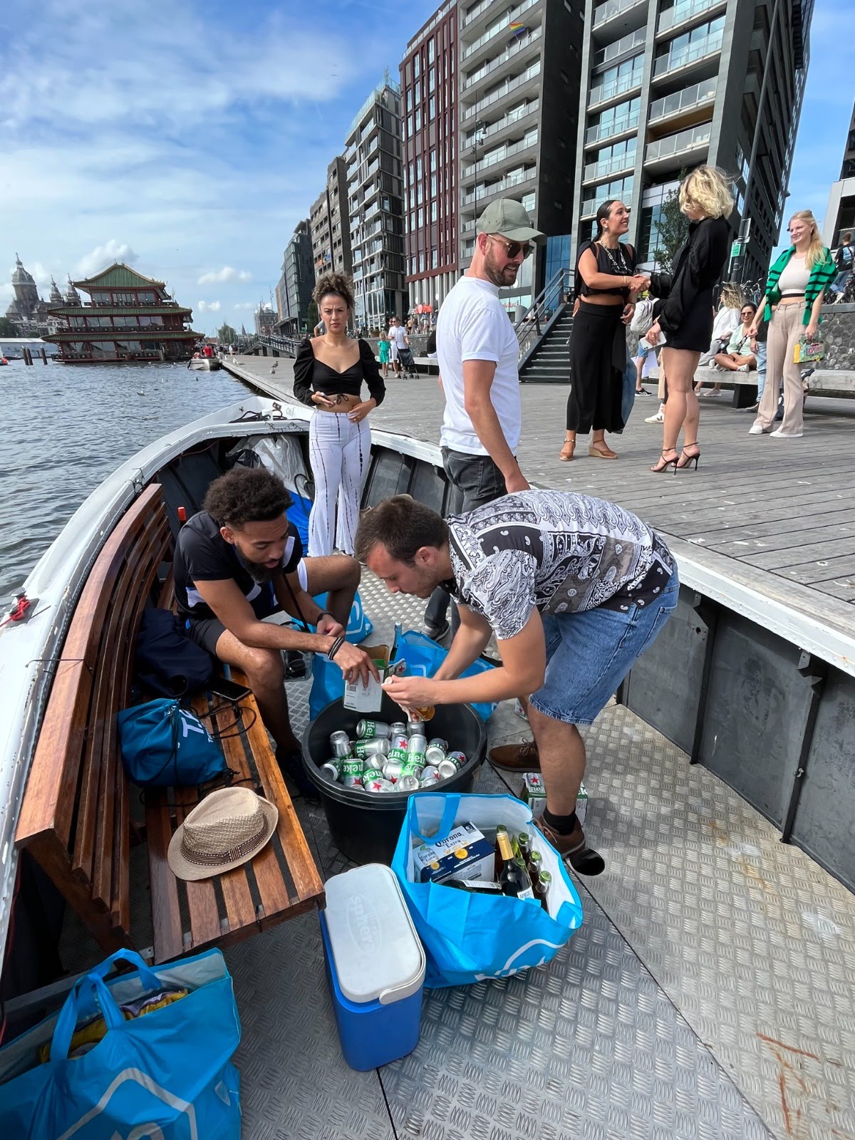 Passengers preparing for a canal boat tour in Amsterdam, stocking up on refreshments against a backdrop of modern riverside architecture and the Sea Palace floating restaurant.