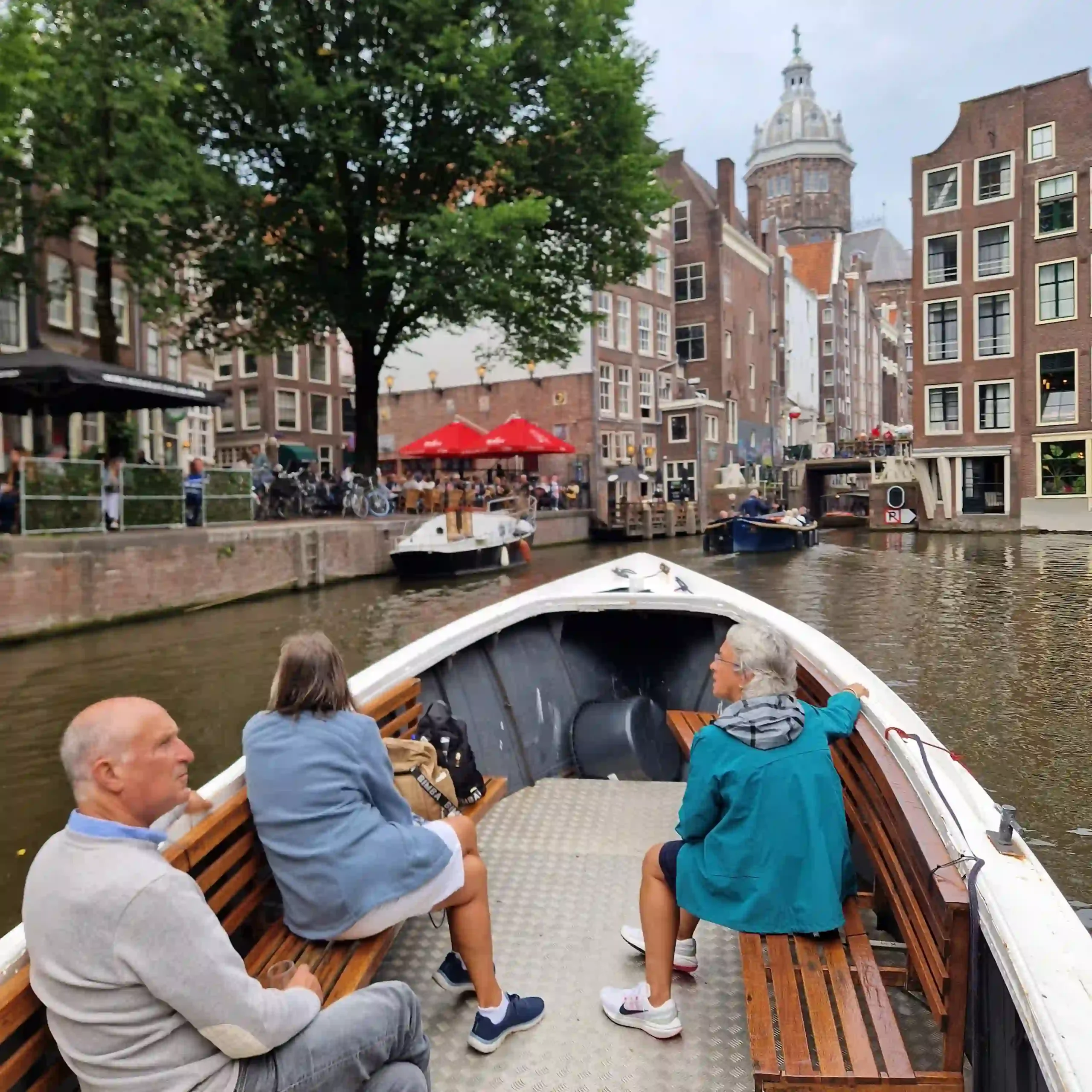 Tourists on a classic Amsterdam canal boat tour gliding past historic gabled buildings and bustling sidewalk cafes